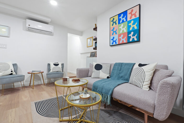 Cozy living room with gray sofa, patterned pillows, glass coffee table, and colorful abstract art on white walls.