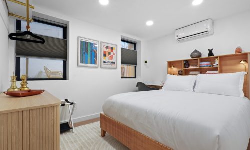 Neatly organized bedroom with a wooden bed frame, white bedding, framed Bauhaus-inspired artwork, and a small desk.
