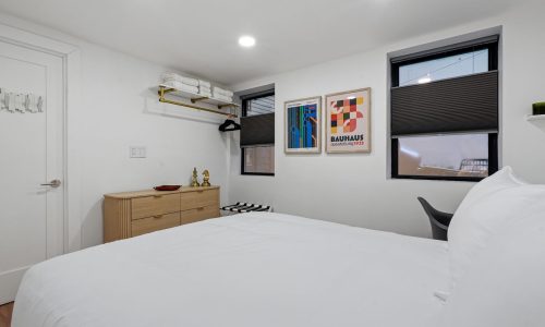 Modern bedroom with white bedding, framed Bauhaus posters, a small desk, and a wooden shelf holding books and decor.