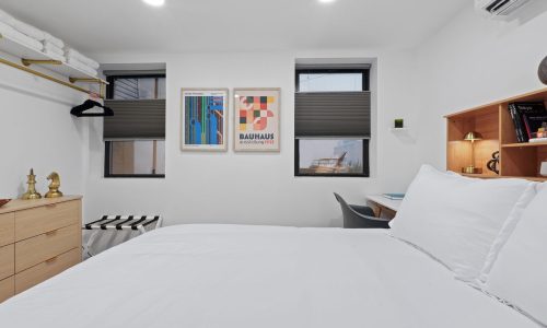 Compact bedroom with a wooden dresser, folding clothes rack, framed Bauhaus poster, towel shelf, and black-and-white striped rug.