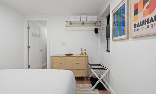 Minimalist bedroom with a wooden dresser, folding clothes rack, framed Bauhaus poster, towel shelf, and gold accents.