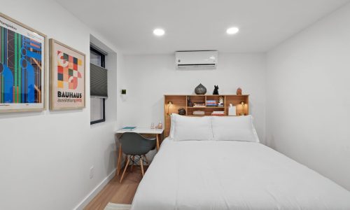 Neatly organized bedroom with white walls, framed Bauhaus-inspired artwork, a small desk, and a wooden shelf above the bed.