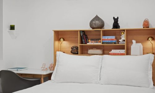 Minimalist bedroom featuring a wooden headboard with built-in shelves, books, decor items, and bedside lamps above a neatly made bed.
