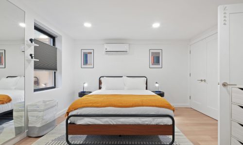 Neatly arranged bedroom with a black-framed bed, white bedding, orange throw blanket, and wooden dresser.