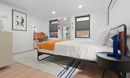 Modern bedroom featuring a black-framed bed with white bedding, orange throw blanket, desk with chair, and framed artwork.