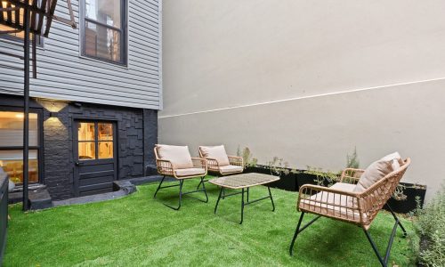 Serene private garden with wicker chairs, metal table, artificial grass, and warm-lit building entrance.