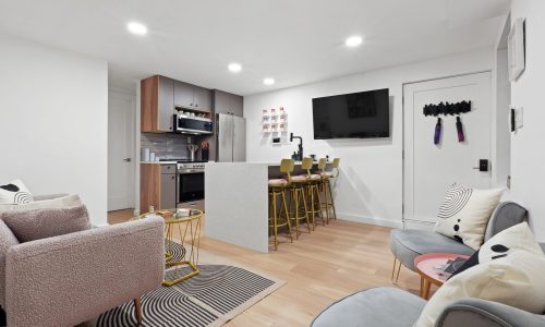 luxury furnished apartments in NYC -- Minimalist open-concept space with kitchen bar, gray sofa, yellow stools, and flat-screen TV.