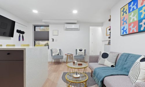 Modern open-concept space with kitchen bar, gray sofa, glass coffee table, and colorful wall art.