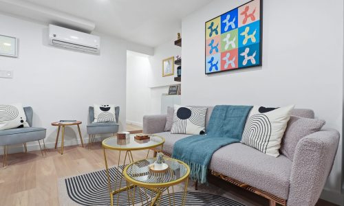 Cozy living room with gray sofa, patterned pillows, glass coffee table, and colorful abstract art on white walls.