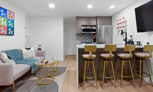 Furnished Studio Apartments in NYC - Modern open-concept apartment with a gray sofa, yellow bar stools, kitchen counter, and colorful wall art.