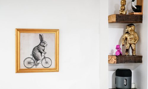 Minimalist wall decor with a gold-framed drawing of a rabbit on a bike, wooden shelves holding a golden astronaut figurine, pink toy, and speaker.