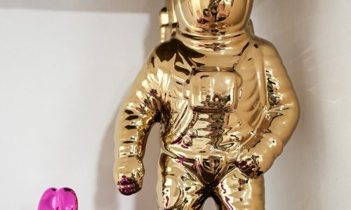 Shiny gold astronaut figurine standing beside a smaller pink metallic dog sculpture on a wooden shelf.