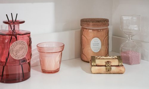 White shelf displaying a red glass bottle, pink candle holder, leather-scented candle, small box, and hourglass.
